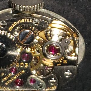 May include: Close-up of a vintage watch movement. The intricate mechanism features gold-colored gears, ruby jewels, and the text "UNADJUSTED" and "17 JEWELS SEVENTEEN". The watch is marked "HAUER WATCH".
