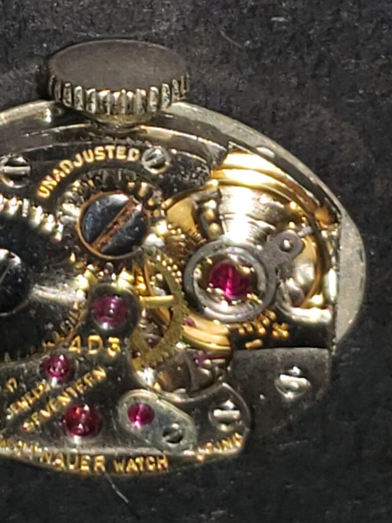 May include: Close-up of a vintage watch movement. The intricate mechanism features gold-colored gears, ruby jewels, and the text "UNADJUSTED" and "17 JEWELS SEVENTEEN". The watch is marked "HAUER WATCH".