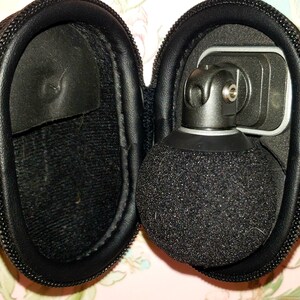 May include: A black microphone with a spherical, fuzzy head is housed in a black, zippered case. The microphone is attached to a metal bracket. The case is open, revealing the microphone and its components. The case is made of a textured material.
