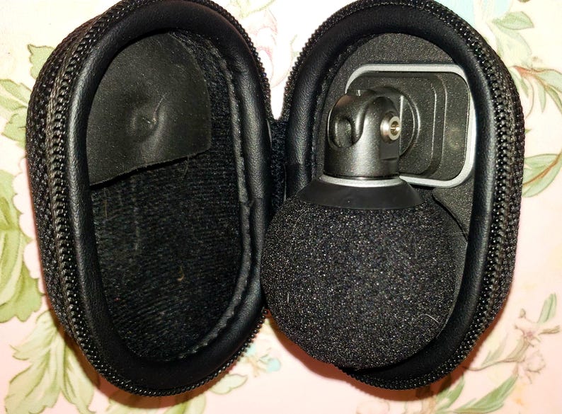 May include: A black microphone with a spherical, fuzzy head is housed in a black, zippered case. The microphone is attached to a metal bracket. The case is open, revealing the microphone and its components. The case is made of a textured material.