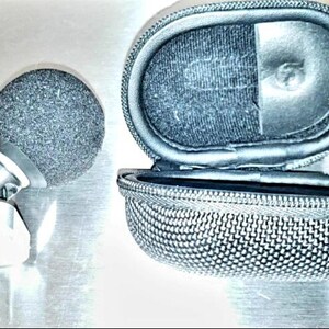 May include: A black microphone with a foam windscreen and a silver-colored mount, next to an open, oval-shaped, zippered case. The case is black with a mesh-like texture on the exterior.