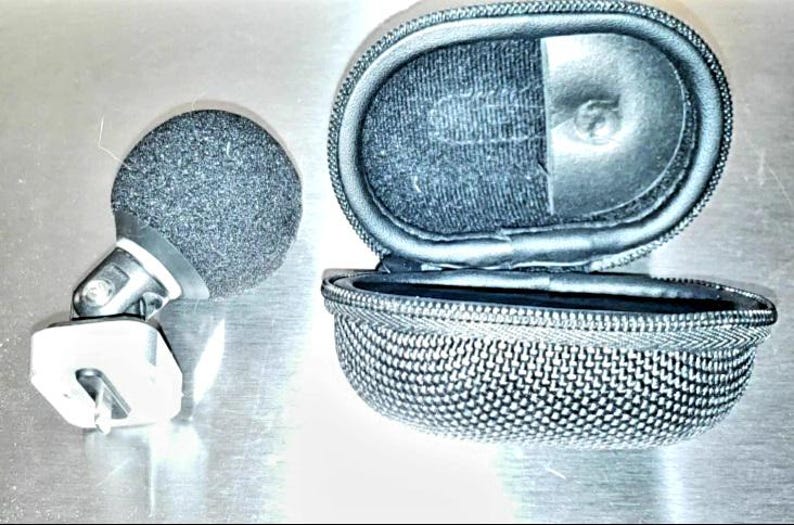 May include: A black microphone with a foam windscreen and a silver-colored mount, next to an open, oval-shaped, zippered case. The case is black with a mesh-like texture on the exterior.