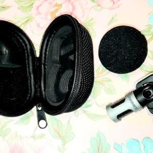 May include: A black zippered carrying case, a black microphone windscreen, and a silver and black microphone mount are displayed on a floral patterned surface. The case is open, revealing its interior. The microphone mount has the brand name "SHURE" printed on it.