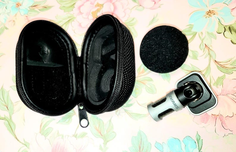 May include: A black zippered carrying case, a black microphone windscreen, and a silver and black microphone mount are displayed on a floral patterned surface. The case is open, revealing its interior. The microphone mount has the brand name "SHURE" printed on it.