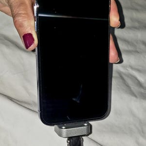 May include: A black smartphone is mounted on a silver and black microphone stand. The phone's screen is black. The stand has a round, black, foam-covered base. The background is a white sheet.