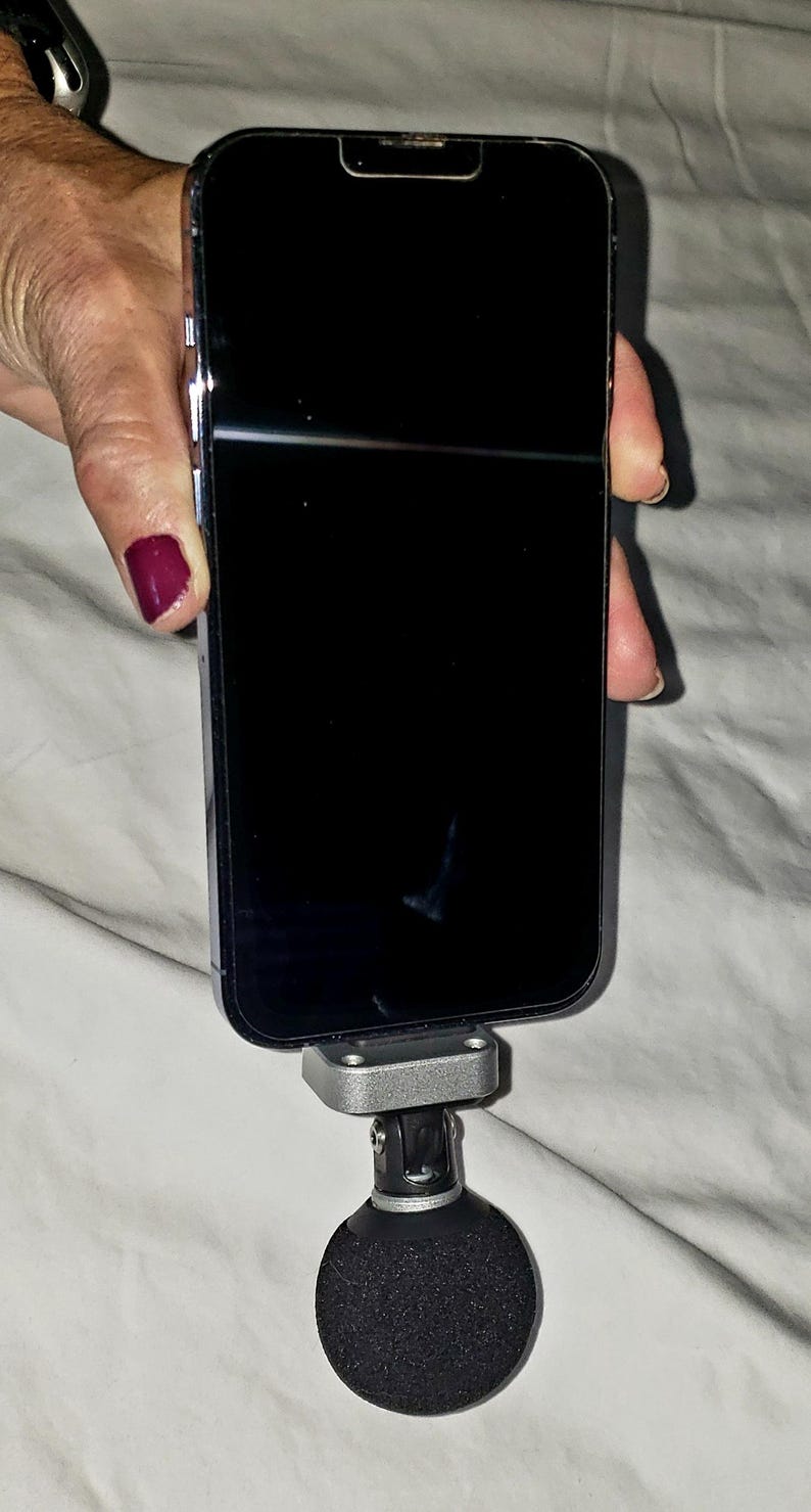 May include: A black smartphone is mounted on a silver and black microphone stand. The phone's screen is black. The stand has a round, black, foam-covered base. The background is a white sheet.