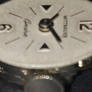 May include: Close-up of a vintage Wittnauer watch face. The watch face is off-white with black hands and markers. The word "Geneve" is printed on the face. The watch has a silver-colored metal casing.