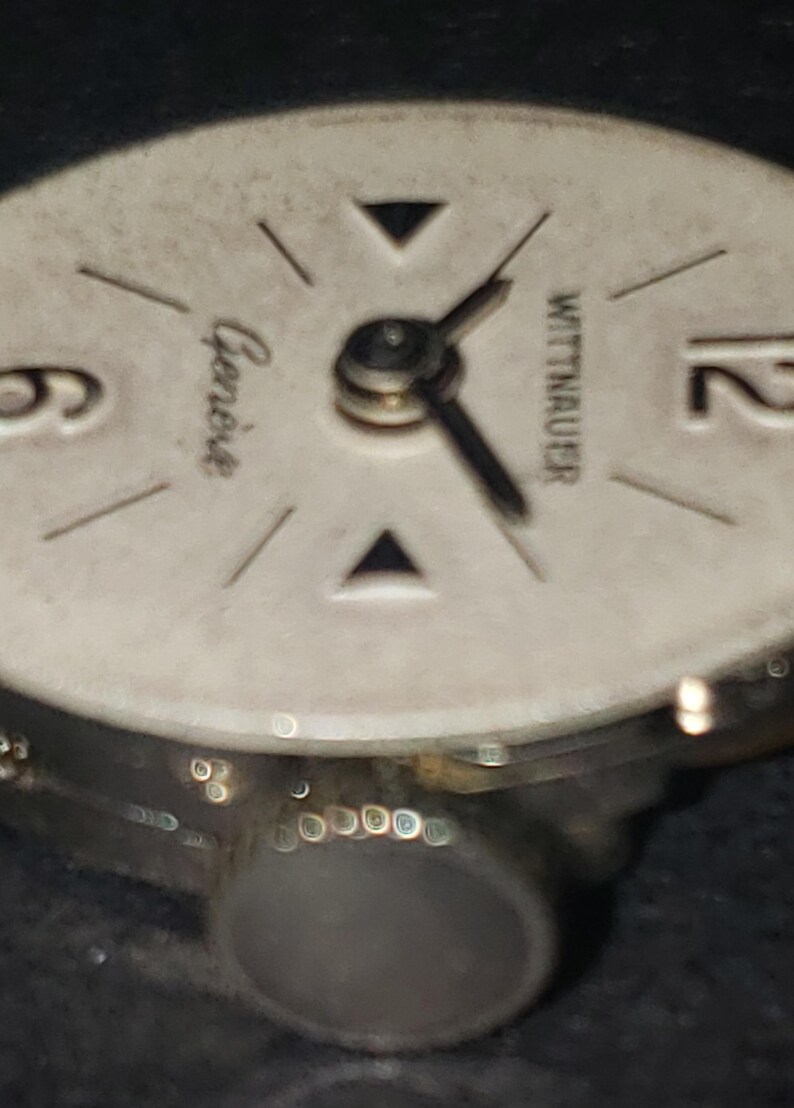 May include: Close-up of a vintage Wittnauer watch face. The watch face is off-white with black hands and markers. The word "Geneve" is printed on the face. The watch has a silver-colored metal casing.