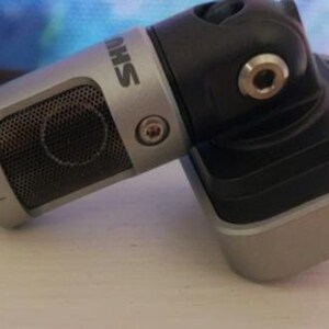 May include: A silver and black microphone with the brand name "SHURE" printed on the side. The microphone has a mesh grill and is attached to a black adjustable stand.