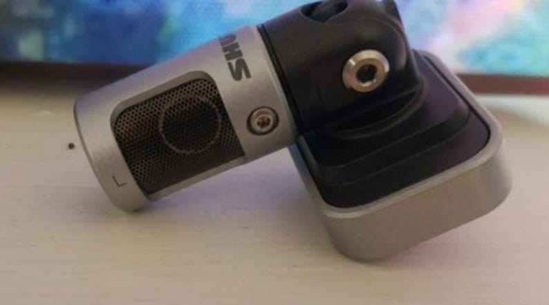 May include: A silver and black microphone with the brand name "SHURE" printed on the side. The microphone has a mesh grill and is attached to a black adjustable stand.