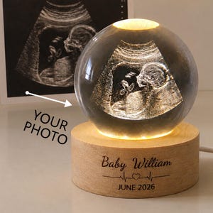 Op de afbeelding: Een bolvormig glazen ornament met een 3D-lasergravure van een echografie, verlicht door een ronde houten voet. De voet is gegraveerd met de tekst Baby William, een hartje met een hartslaglijn en de datum JUNE 2026.
