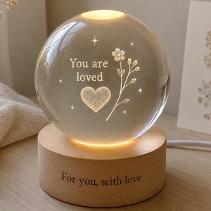 May include: A clear glass sphere lamp resting on a cylindrical light-coloured wooden base. The sphere features an internal laser-etched design of a heart, a flower, and the text "You are loved." The wooden base is engraved with "For you, with love."