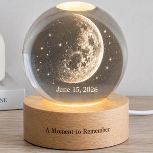 May include: A spherical glass orb featuring an internal laser-etched moon and star design, illuminated by a circular light-colored wooden base. The glass displays the text June 15, 2026, while the wooden base is engraved with A Moment to Remember.