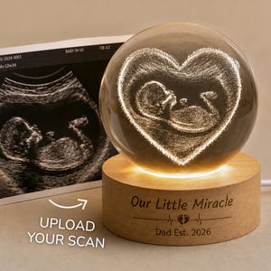 Op de afbeelding: Een heldere glazen bol met een lasergegraveerde foetus in een hartvorm, gemonteerd op een ronde houten voet. De voet is gegraveerd met de tekst "Our Little Miracle", een hartslaglijn met babyvoetjes en "Dad Est. 2026".