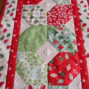 May include: A red, green, and white quilted table runner with a Christmas theme. The runner features a variety of holiday patterns, including snowflakes, Christmas trees, and lights.