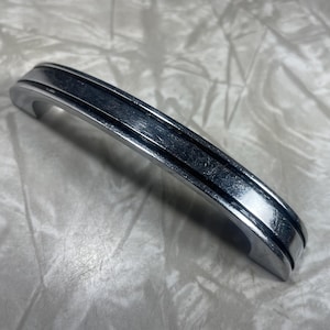 May include: A silver-toned, curved cabinet pull with a grooved center detail. The handle is arched and has a smooth, reflective surface. The hardware is designed for furniture and has a classic, understated aesthetic.