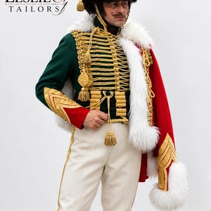 Puede incluir: Una chaqueta de estilo militar verde oscuro con adornos de cordón dorado, botones dorados y borlas, usada con pantalones blancos y una capa roja con ribete de piel sintética blanca. El texto LESLIE & TAILORS es visible en la esquina superior izquierda.