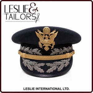 May include: A dark navy blue military service cap featuring a prominent gold-toned eagle emblem on the front. The cap includes a gold chin strap and silver-colored embroidered oak leaf detailing on the band and visor. Text reads LESLIE & TAILORS.