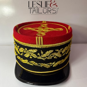 Puede incluir: Sombrero de estilo militar rojo y negro con bordados dorados. El sombrero presenta un borde negro y una parte superior roja con adornos dorados. El texto "LESLIE & TAILORS" es visible en el fondo.