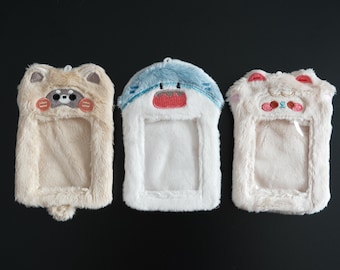 May include: Three plush phone cases with animal designs. The cases are rectangular with a clear window for the phone screen. The first case is tan with a raccoon face, the second is white and blue with a shark face, and the third is white with a bear face.