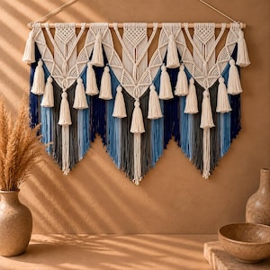 May include: A large macrame wall hanging suspended from a wooden dowel, featuring intricate off-white knotted patterns and numerous hanging tassels. The lower section consists of layered vertical fringe in shades of navy blue, light blue, and grey.