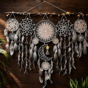 May include: A large wall hanging featuring multiple circular crochet dreamcatchers suspended from a dark wooden branch. The design includes grey feathers, a crescent moon shape, a small gold star, and warm white string lights wrapped around the branch.