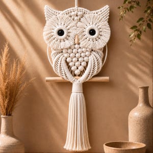 May include: A cream-coloured macrame wall hanging shaped like an owl, featuring intricate knotted patterns, two large circular eye details with black centres, a wooden perch, and a long, thick tassel hanging from the bottom centre of the owl figure.