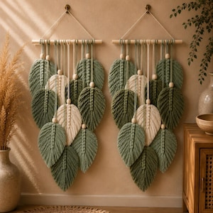 Op de afbeelding: Twee identieke macramé wandkleden met meerdere bladvormige vezelelementen in saliegroen en crème. Elk stuk hangt aan een horizontale houten stok met een wit koord en is versierd met natuurlijke houten kralen.