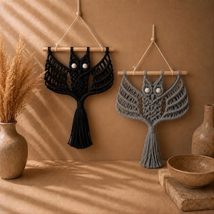 May include: Two macrame wall hangings shaped like owls, one in black and one in gray. Each owl is crafted from knotted cord, features two white circular eyes, and hangs from a horizontal wooden dowel suspended by a thin cord loop.