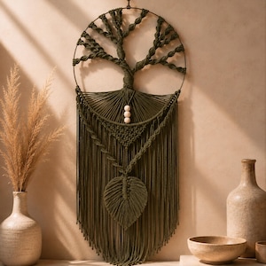 Op de afbeelding: Een ronde macramé wandhanger in olijfgroen met een levensboomontwerp in een metalen ring. Het werk bevat ingewikkelde geknoopte takken, drie lichtgekleurde houten kralen, een driehoekig geweven gedeelte en een centraal bladmotief.