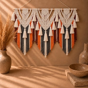 Peut inclure: Une tenture murale en macramé composée de cordes couleur crème disposées en motifs géométriques complexes avec de multiples pompons. Le design intègre des brins verticaux dans des tons orange, marron foncé et gris, suspendus à une tige en bois.