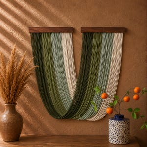 May include: A wall hanging featuring two dark wood horizontal bars from which long, thin, vertical strands of fibre drape in a U-shape. The strands transition in colour from deep forest green to olive, sage, and cream, creating a layered gradient.