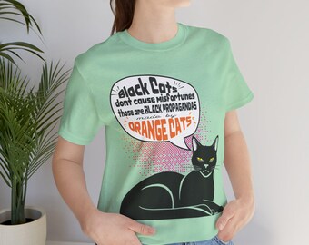 May include: A pale green t-shirt with a black cat illustration and text in a speech bubble. The text says "Black Cats don't cause misfortunes those are BLACK PROPAGANDAS made by ORANGE CATS."