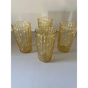 May include: Four amber-colored drinking glasses. Each glass has a textured, vertical pattern. The glasses are cylindrical with a slightly flared top. The glasses are made of glass and are suitable for serving beverages.
