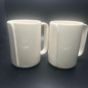 May include: Two off-white ceramic mugs with a simple, geometric design. Each mug has a handle and a slightly flared rim. The mugs are set against a dark background, highlighting their clean lines and neutral color. Ideal for coffee or tea.