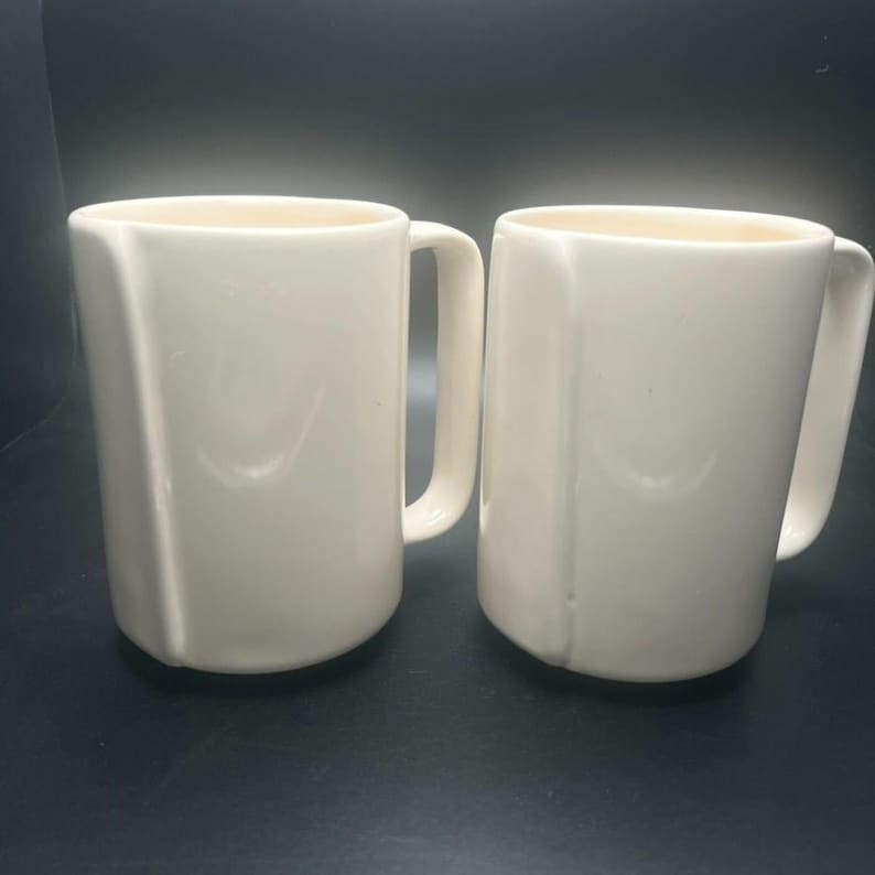 May include: Two off-white ceramic mugs with a simple, geometric design. Each mug has a handle and a slightly flared rim. The mugs are set against a dark background, highlighting their clean lines and neutral color. Ideal for coffee or tea.