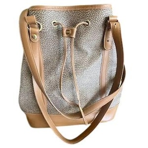 May include: A tan and brown bucket bag with a drawstring closure. The bag features a textured pattern and tan leather straps and trim. The bag has a single shoulder strap and a smaller strap for carrying.