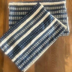 May include: Two blue and white patterned throw pillows. The pillows have a striped design with white and blue vertical stripes. The top pillow is folded in half. The pillows are on a wooden surface.