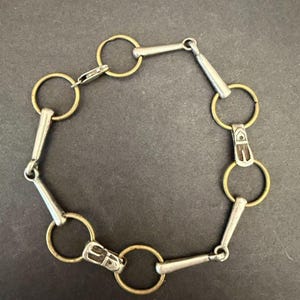 May include: A bracelet constructed from alternating circular brass rings and silver-toned metal bars. The design features small rectangular silver-toned connectors with engraved detailing that link the rings and bars together in a continuous chain.