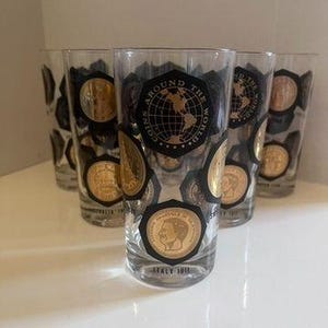 May include: Set of six vintage highball glasses. Each glass features a black and gold design with coin illustrations and the text "AROUND THE WORLD COINS". The glasses have a clear base and are ideal for serving beverages.