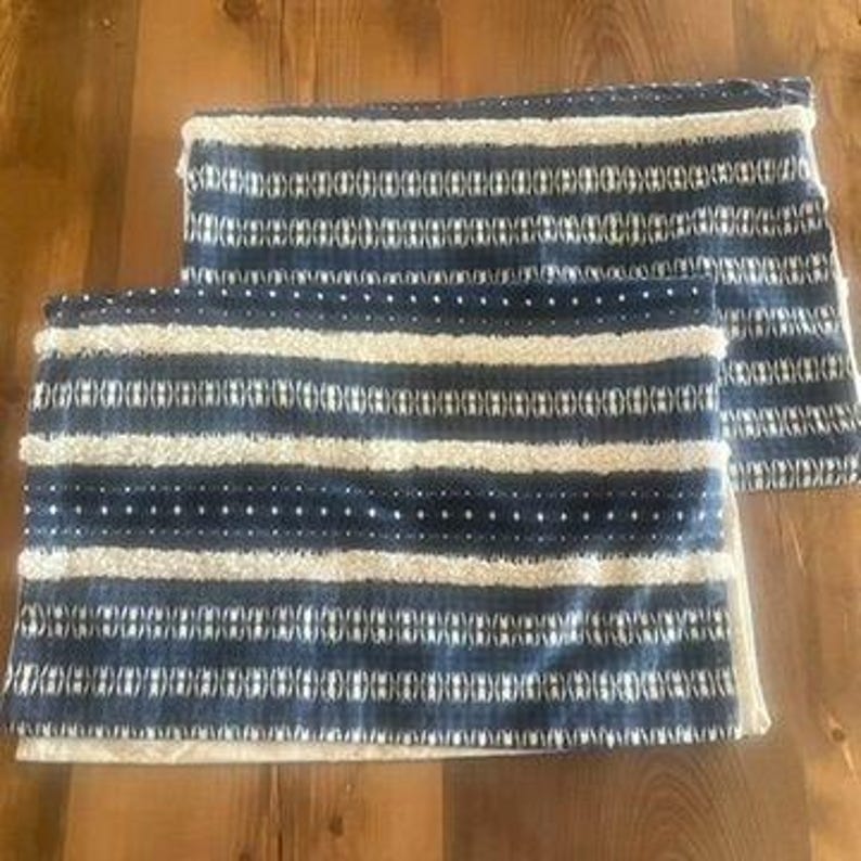 May include: Two rectangular throw pillows with a blue and white striped pattern. The pillows have a textured appearance with a combination of horizontal stripes and dotted designs. The pillows are placed on a wooden surface.