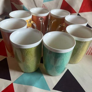 Op de afbeelding: Een verzameling van acht keramische mokken met een witte binnenkant en een textuurrijke, kleurrijke buitenkant. De mokken hebben doorzichtige glazen handvatten en zijn gerangschikt op een patroon. Kleuren zijn oranje, groen, geel en teal.