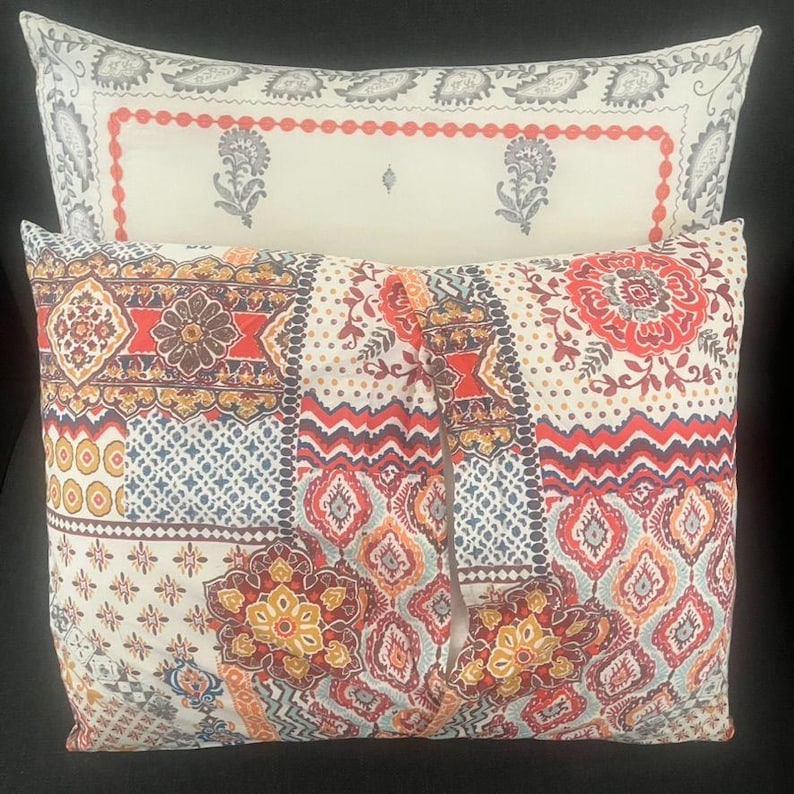 May include: Two decorative throw pillows with intricate patterns. The top pillow has a cream background with a red border and floral designs. The bottom pillow features a mix of floral and geometric patterns in red, orange, blue, and yellow.