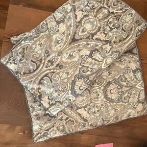 Może przedstawiać: Prostokątna poszewka na poduszkę z detalicznym wzorem paisley w odcieniach szarości, beżu i delikatnego błękitu. Tkanina ma pikowaną fakturę z widocznymi liniami przeszyć na całym wzorze, wykończona lamówką na krawędziach.