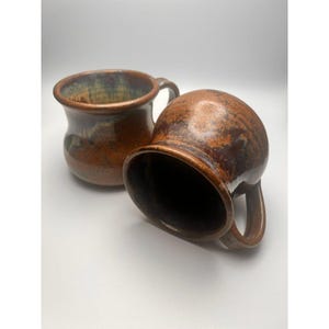 May include: Two handmade ceramic mugs with a rustic, earthy aesthetic. The mugs are a rich brown color with subtle variations and a textured surface. Each mug has a rounded body, a sturdy handle, and a wide opening.