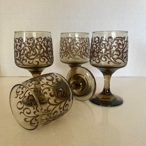 Puede incluir: Juego de cuatro copas de vino vintage de color ámbar. Cada copa presenta un diseño ornamental marrón y arremolinado alrededor del cuenco. Una copa está tumbada de lado, mientras que las otras están de pie. Las copas tienen un tallo y una base redonda.