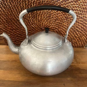 May include: A vintage aluminium kettle with a black handle and spout. The kettle has a rounded body and a lid with a small black knob. The handle is curved and attached to the sides. It sits on a wooden surface.