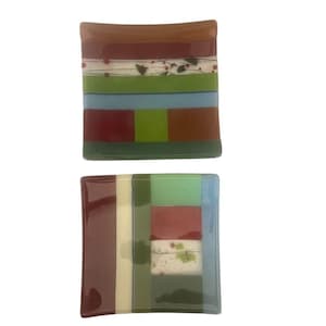 Può includere: Due piatti quadrati di vetro con disegni geometrici colorati. Il piatto superiore presenta strisce e blocchi marroni, rossi, verdi e blu. Il piatto inferiore ha colori simili, disposti in motivi verticali e orizzontali.