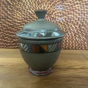 May include: A ceramic jar with a lid, featuring a dark green base and lid with a geometric pattern in shades of blue, orange, and white. The jar sits on a small, round base. The background includes a woven wicker texture.
