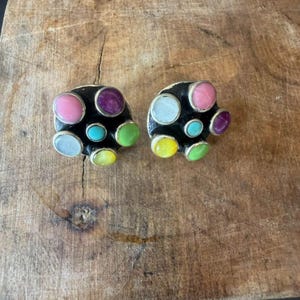 May include: A pair of circular earrings featuring a black base set with multiple small, round, multicolored cabochons in shades of pink, purple, turquoise, yellow, light green, and white, each framed in a thin silver-toned metal border.